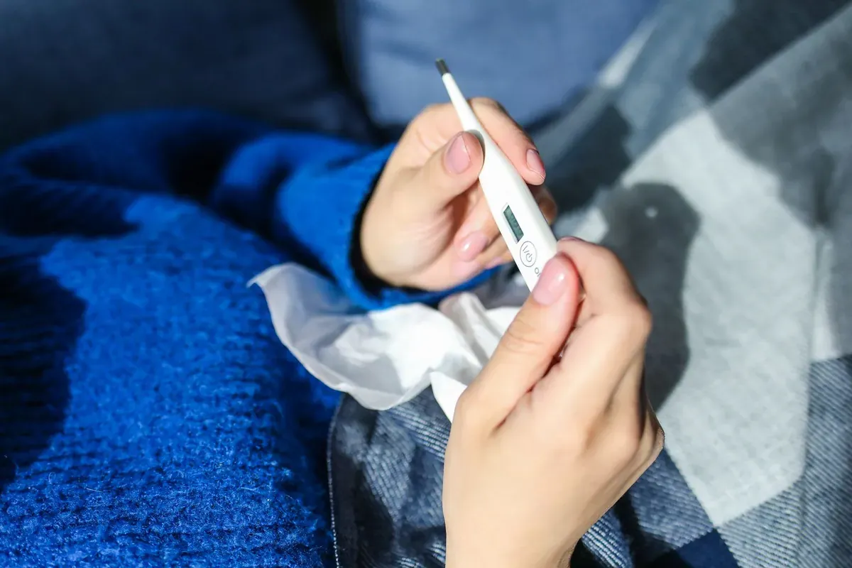 Eine Person hält ein Fieberthermometer und ruht unter einer Decke